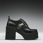 A black leather platform clog shoe with a chunky black sole and heel, featuring a decorative silver skeleton hand ornament on the side, set against a dark gray gradient background.