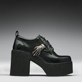 A black leather platform clog shoe with a chunky black sole and heel, featuring a decorative silver skeleton hand ornament on the side, set against a dark gray gradient background.