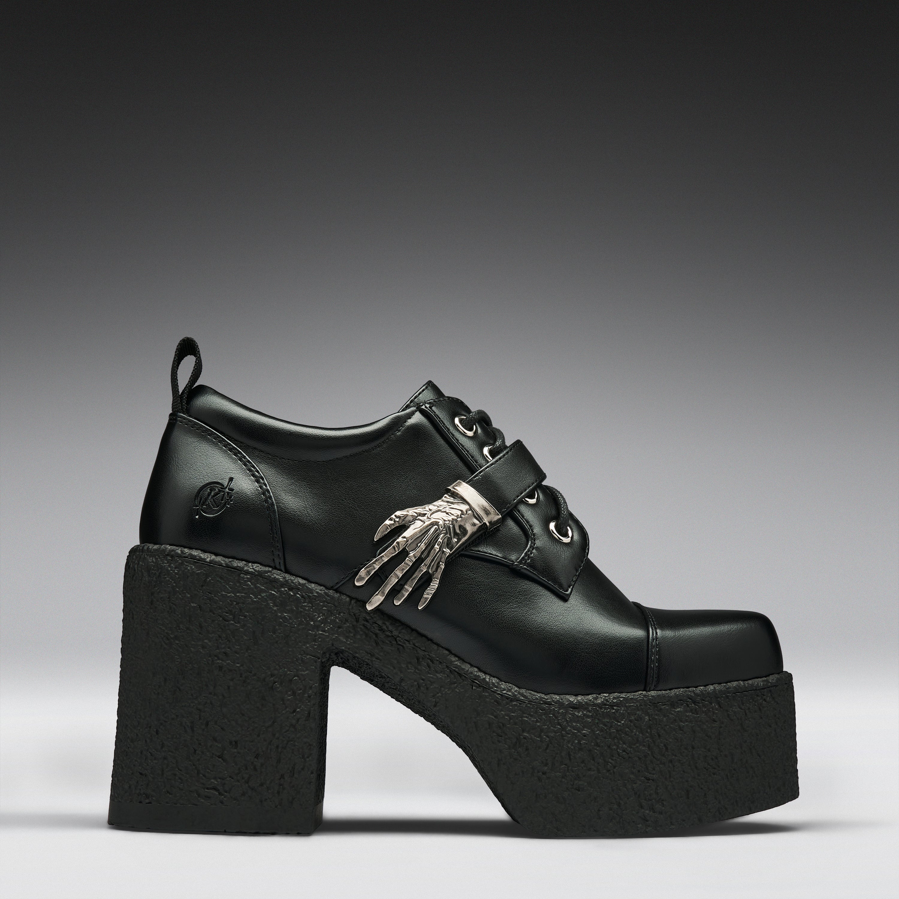 A black leather platform clog shoe with a chunky black sole and heel, featuring a decorative silver skeleton hand ornament on the side, set against a dark gray gradient background.