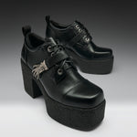 A pair of black leather platform lace-up shoes with chunky block heels, decorative straps, and silver hardware, photographed on a dark-to-light gray gradient studio background.