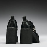 Black leather platform ankle boots with chunky textured block heels and silver chain details, shown from the back on a dark gray gradient background.