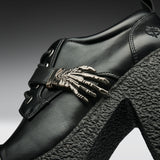 Close-up of a black leather high-heeled shoe featuring a prominent metallic skeleton hand ornament on the side strap, a chunky black textured platform heel, and a small embroidered logo near the heel.