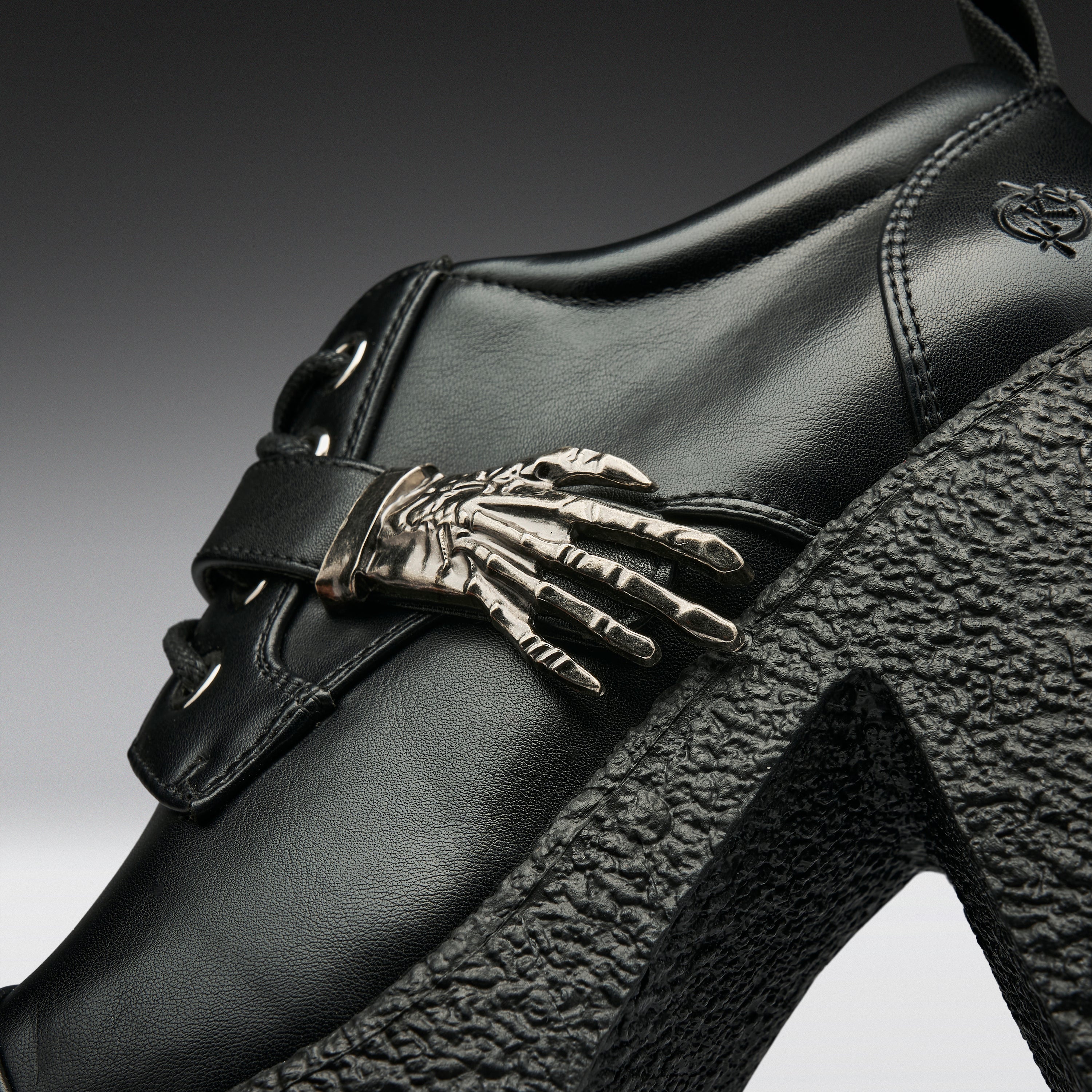 Close-up of a black leather high-heeled shoe featuring a prominent metallic skeleton hand ornament on the side strap, a chunky black textured platform heel, and a small embroidered logo near the heel.