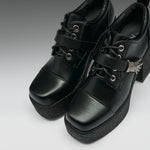 A pair of black platform lace-up ankle boots with chunky black soles, silver eyelets, and a small silver decorative accent on the side, shown against a gray gradient background.