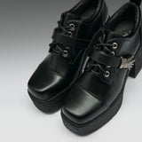 A pair of black platform lace-up ankle boots with chunky black soles, silver eyelets, and a small silver decorative accent on the side, shown against a gray gradient background.