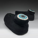 A pair of black plush slipper boots with circular eye motifs on the back, photographed on a dark-to-light gray gradient studio background.