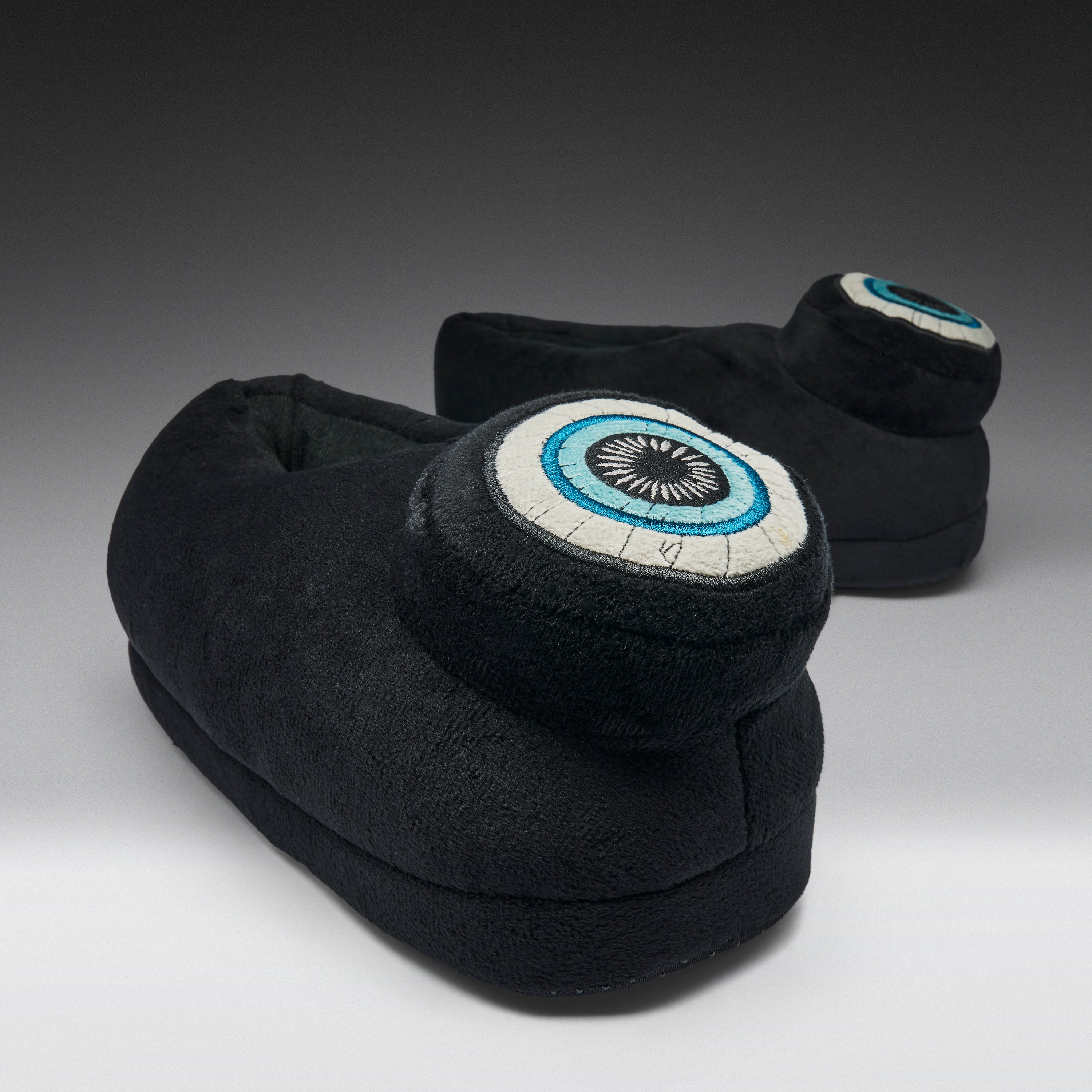 A pair of black plush slipper boots with circular eye motifs on the back, photographed on a dark-to-light gray gradient studio background.