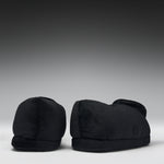 A pair of black plush slippers shown from the back against a dark gray gradient studio background.