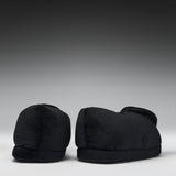 A pair of black plush slippers shown from the back against a dark gray gradient studio background.