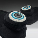 A pair of black plush slippers with embroidered circular eye designs on the back, shown on a dark gray to white studio background.