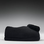 A black fleece-covered footrest or cushion with a curved shape and a small circular white accent on top, photographed against a dark gray to light gray gradient studio background.
