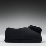 A black fleece-covered footrest or cushion with a curved shape and a small circular white accent on top, photographed against a dark gray to light gray gradient studio background.