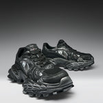 A pair of black and dark gray chunky athletic sneakers with sculpted, textured soles and metallic silver graphic details, displayed on a dark-to-light gray gradient studio background.