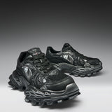 A pair of black and dark gray chunky athletic sneakers with sculpted, textured soles and metallic silver graphic details, displayed on a dark-to-light gray gradient studio background.