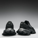 A pair of black chunky sneakers shown from the rear, featuring textured black soles and a circular logo on the heel, set against a dark gray gradient studio background.