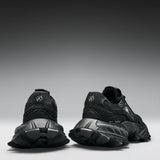 A pair of black chunky sneakers shown from the rear, featuring textured black soles and a circular logo on the heel, set against a dark gray gradient studio background.