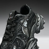 A close-up studio shot of a black chunky sneaker with sculptural layered soles, mesh side panels, and graphic face details in white and gray, photographed against a dark gray gradient background.