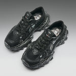A pair of black chunky sneakers with textured, sculptural soles and mesh panels, photographed on a clean gray gradient background.