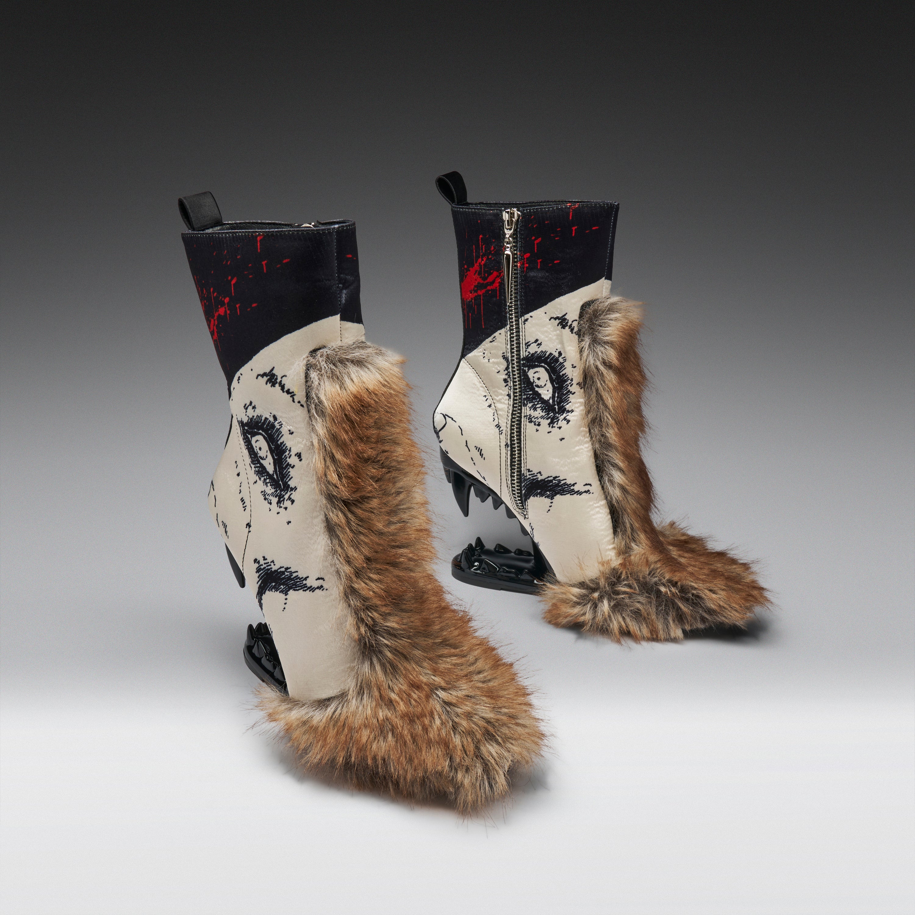A pair of avant-garde ankle boots with beige and black printed panels, red splatter details, and thick brown fur trim, shown on a gray gradient studio background.
