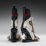 A pair of high-heeled boots shown from the back, featuring glossy black wedge heels with sharp teeth-like details, cream-colored panels with black graphic print, red paint splatter accents, and brown fur trim, photographed on a gray gradient studio background.