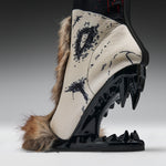 A single high-heeled boot shown in side profile on a studio background. The boot has a cream-colored upper with black graphic artwork, brown fur trim, and a glossy black sculptural heel and sole with sharp spike details.