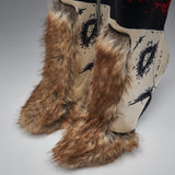 Close-up of a pair of western-style boots with long brown faux-fur leg warmers and foot covers, cream shafts with black eye-like graphics, and a black top section with red splatter details.
