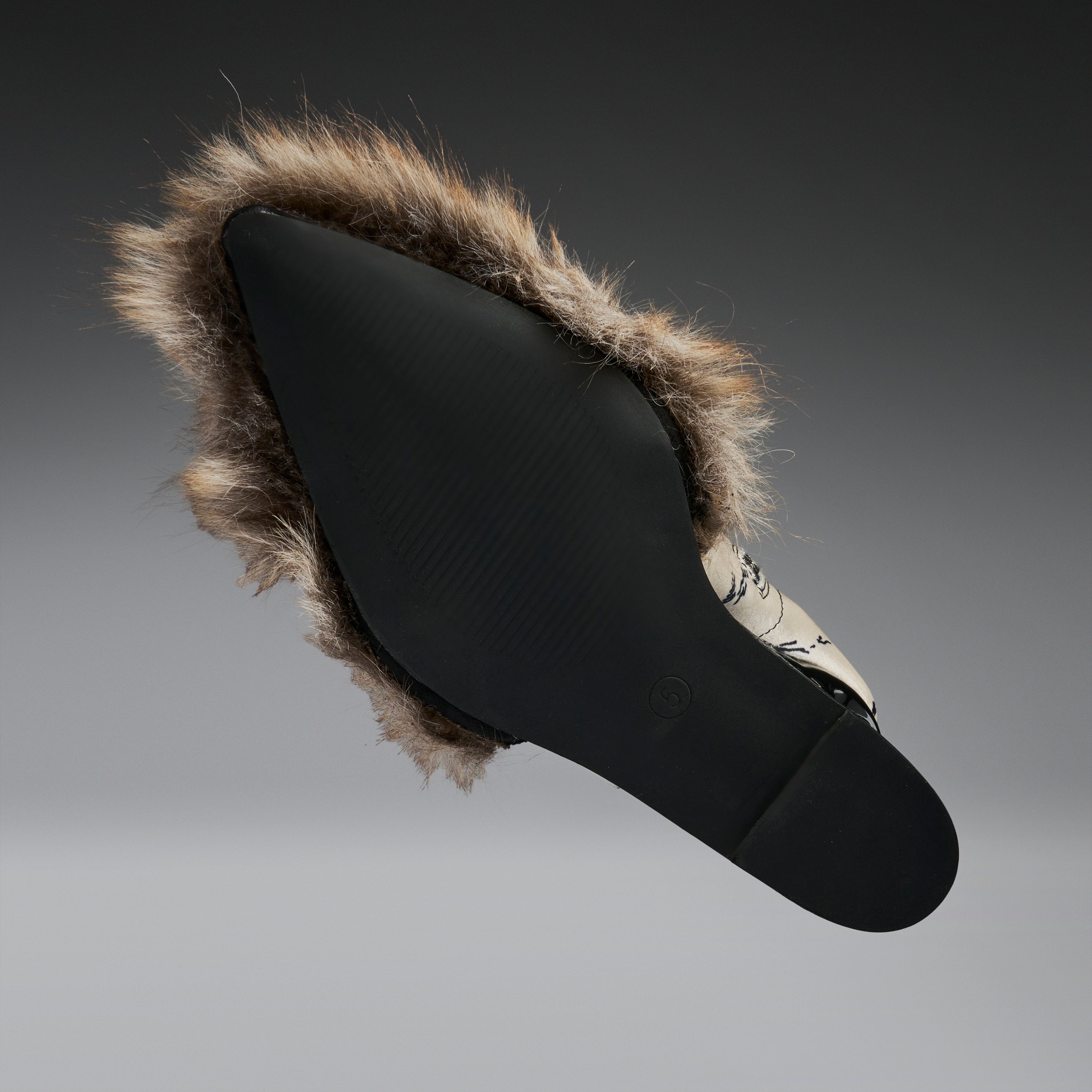 A black slip-on shoe with a brown and beige faux-fur trim, shown from a side profile against a dark gray gradient background.