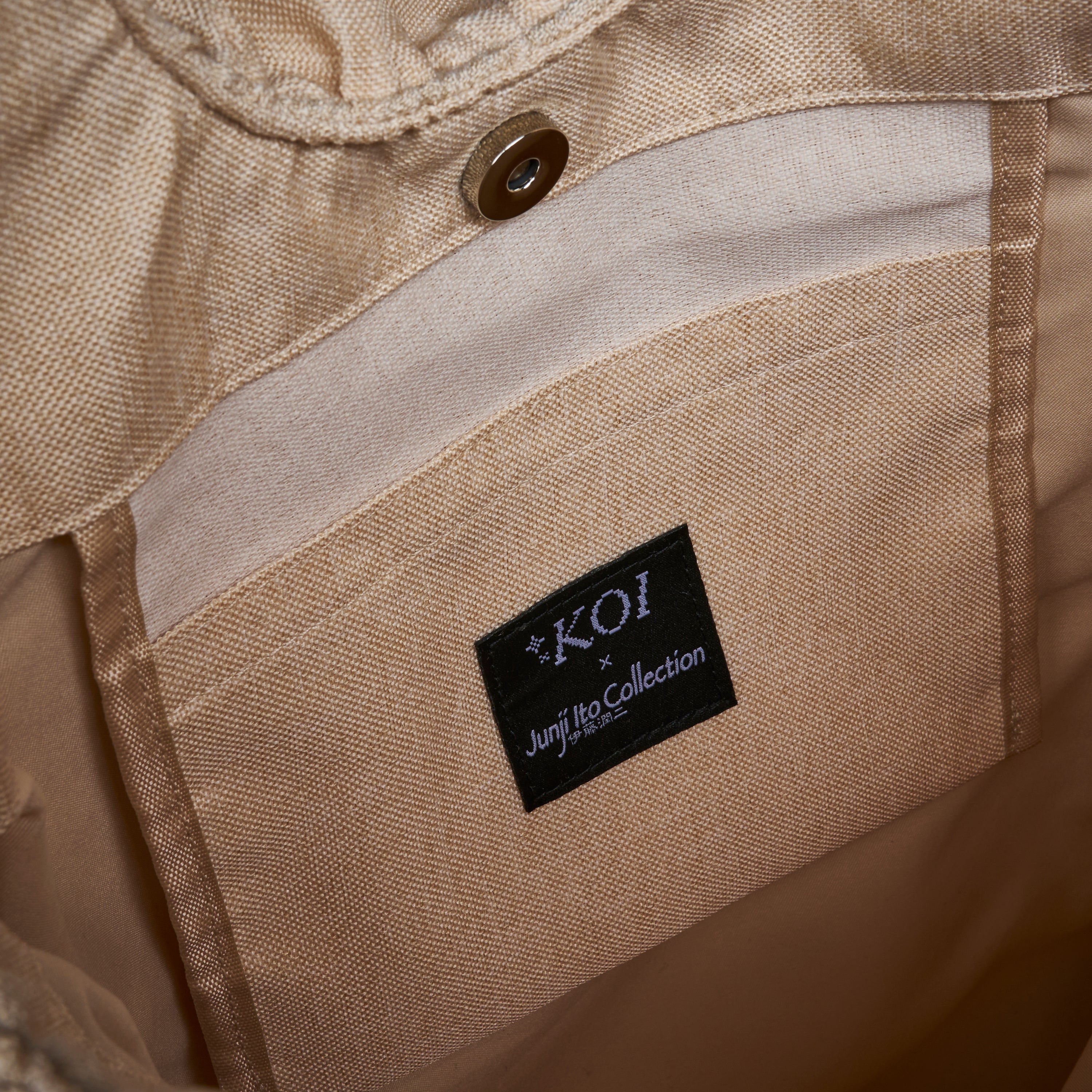 Close-up of the interior lining of a beige denim garment, showing a black brand label that reads 'KOI x Junji Collection' sewn onto a tan fabric pocket.