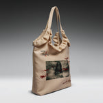 A beige canvas tote bag with ruffled trim and dual shoulder straps, featuring a horror-themed graphic print with a dark figure and red splatter details, and the text 'KOI' on the front, shown against a dark-to-light gray gradient background.