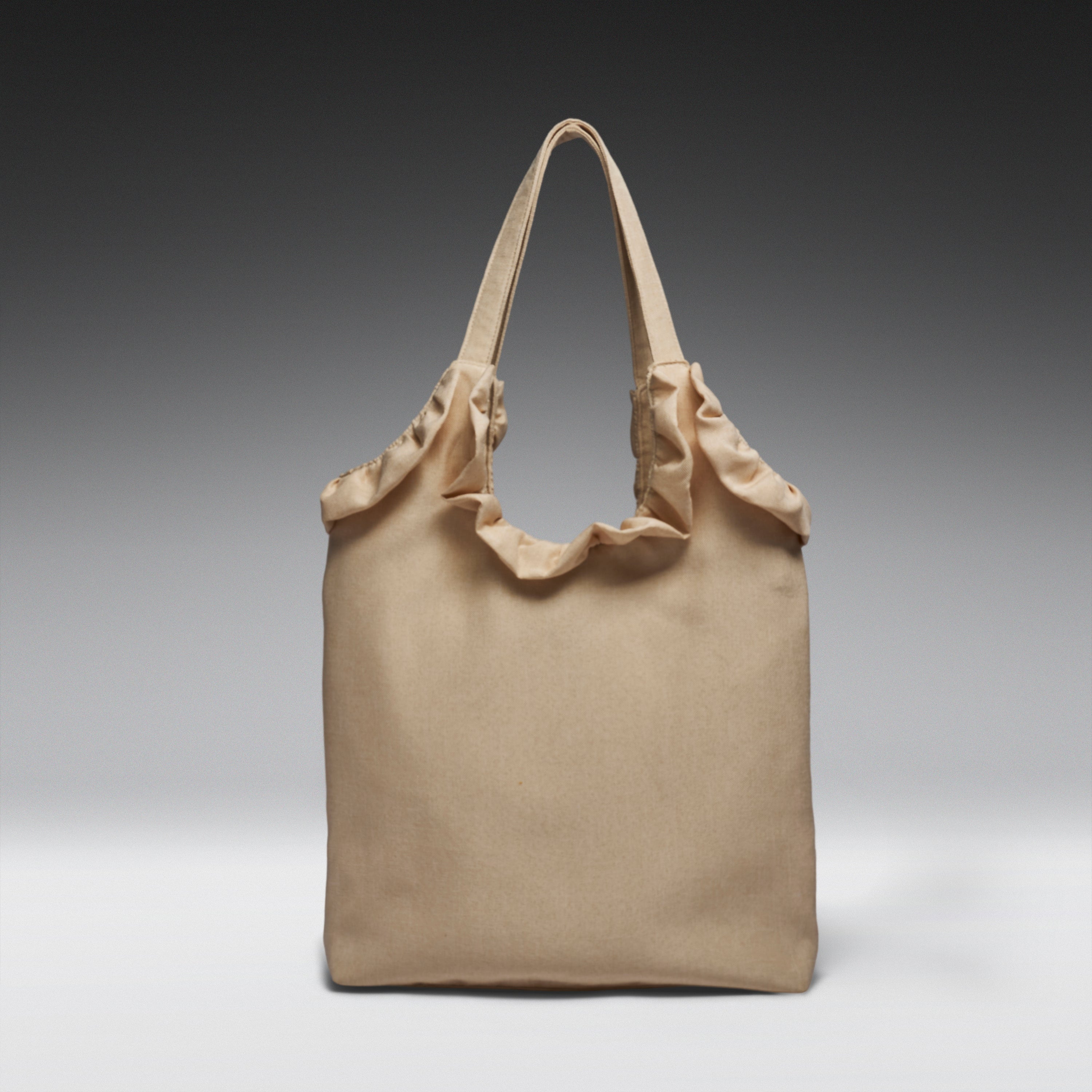 A beige canvas tote bag with gathered fabric trim along the top and dual shoulder straps, photographed against a dark-to-light gray gradient studio background.