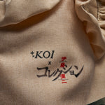 Close-up of a beige woven fabric surface with embroidered text reading “*KOI x Junji Ito” in black and red.
