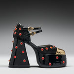 A single black patent leather platform sandal with a chunky block heel, embellished with red rhinestones and gold-tone hardware, shown against a dark gray gradient background.