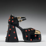A single black patent leather platform sandal with a chunky block heel, embellished with red rhinestones and gold-tone hardware, shown against a dark gray gradient background.