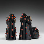 A pair of black platform high heels with glossy patent block heels, fuzzy black uppers, and numerous red rhinestone studs with gold-toned settings, shown from the back against a dark-to-light gray gradient studio background.