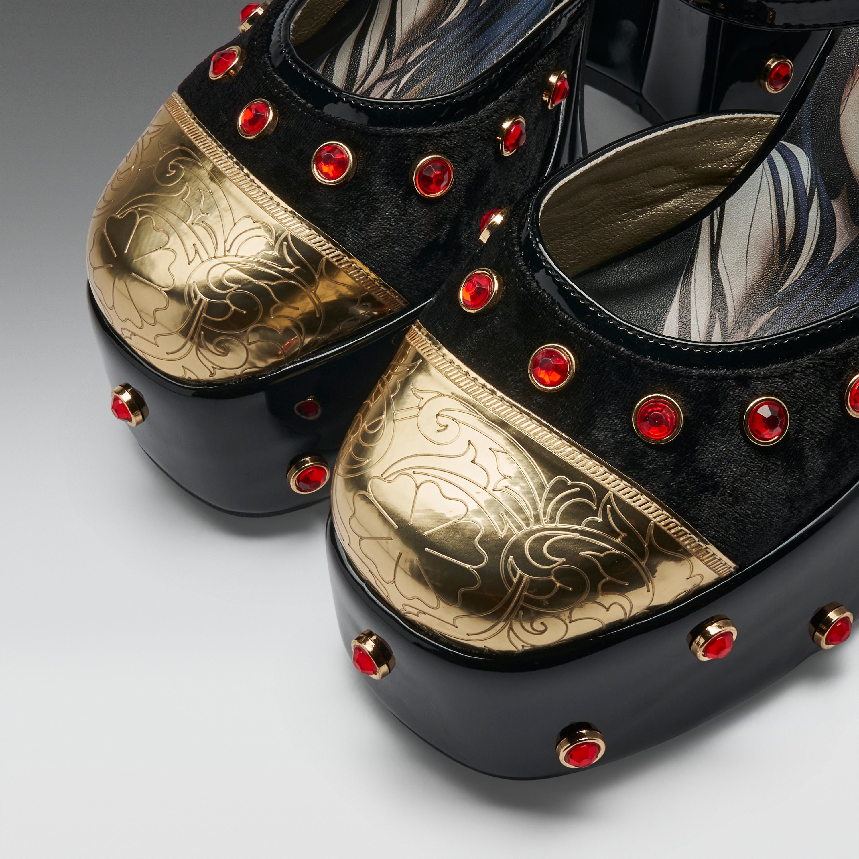 A close-up of a pair of platform Mary Jane shoes featuring black velvet uppers, metallic gold floral-embossed toe caps, black platform soles, and red rhinestone embellishments.