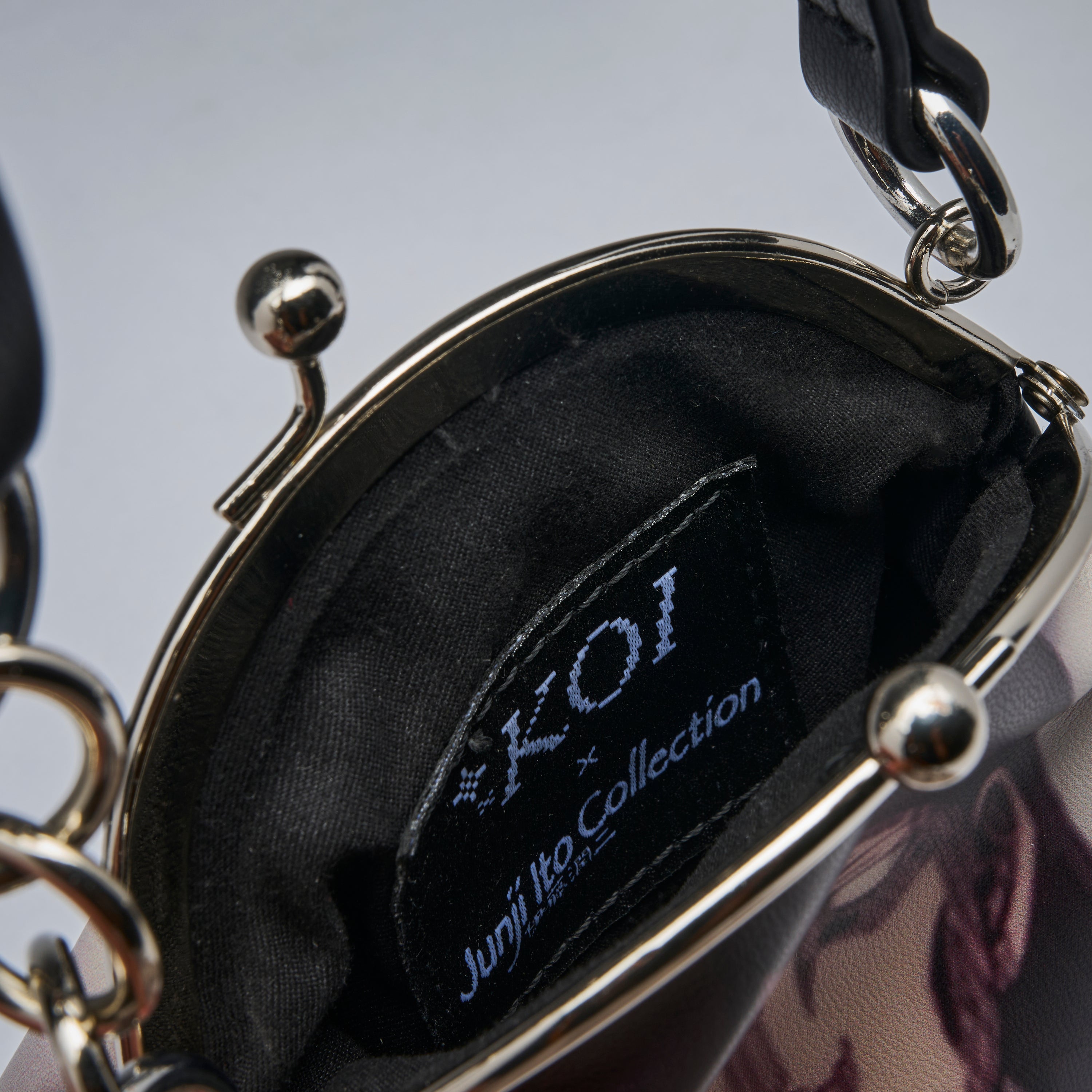 Close-up of the interior of a handbag showing a black fabric lining with a visible brand patch that reads 'Koi x Junji Ito Collection'. The bag has a silver-tone metal frame and chain details, with a dark patterned exterior partially visible at the bottom right.