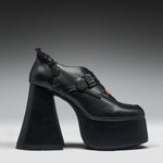 A single black platform high-heel shoe with a chunky sculptural block heel, a buckle strap across the instep, and a small orange accent near the toe, photographed against a dark-to-light gray gradient studio background.
