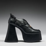 A single black platform high-heel shoe with a chunky sculptural block heel, a buckle strap across the instep, and a small orange accent near the toe, photographed against a dark-to-light gray gradient studio background.