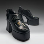 A pair of black platform mule heels with ruffled trim, shown on a clean gray studio background. The shoes feature a chunky block heel, a thick platform sole, a buckle strap across the top, and a glossy finish with a small printed graphic on the toe area.