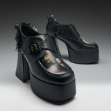 A pair of black platform mule heels with ruffled trim, shown on a clean gray studio background. The shoes feature a chunky block heel, a thick platform sole, a buckle strap across the top, and a glossy finish with a small printed graphic on the toe area.