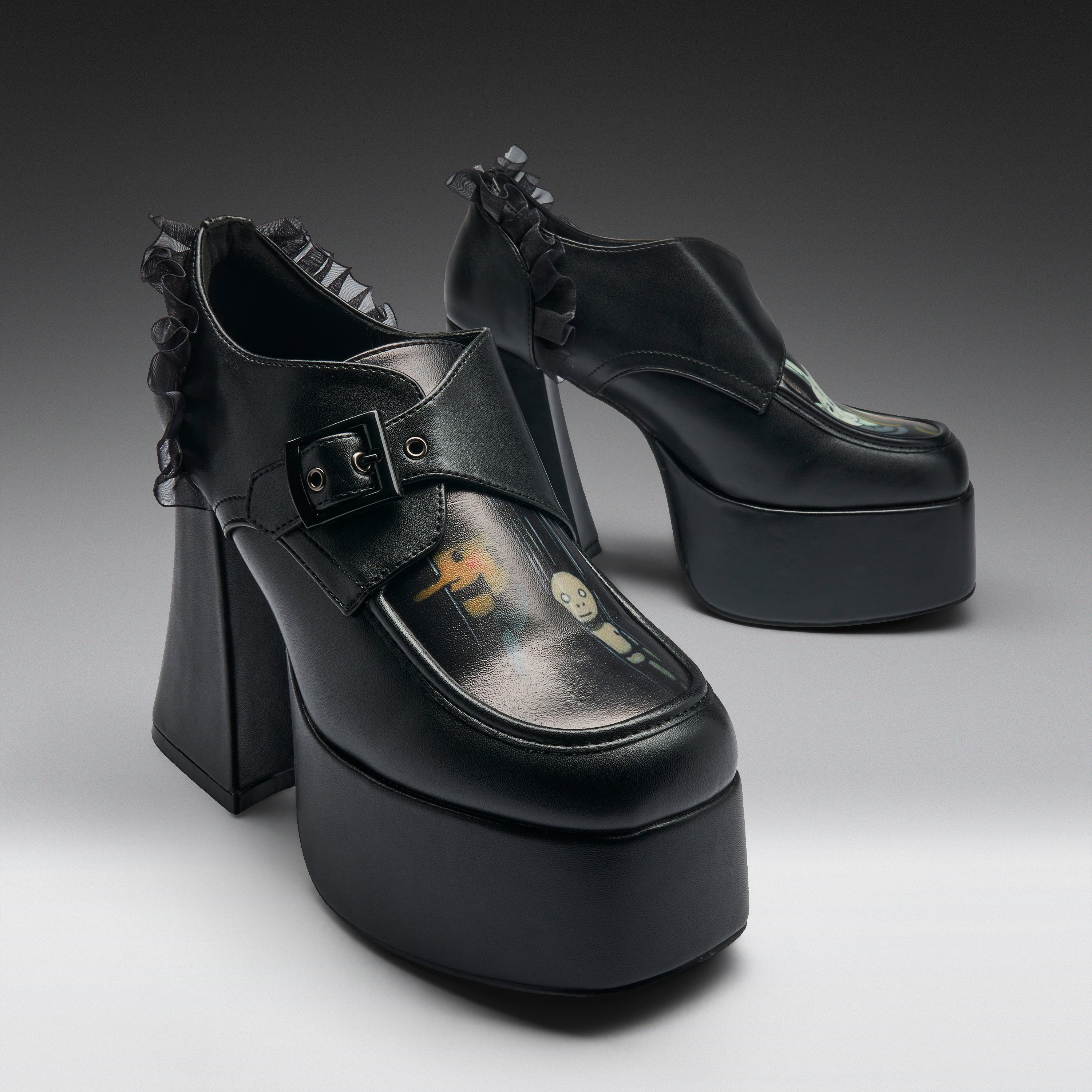 A pair of black platform mule heels with ruffled trim, shown on a clean gray studio background. The shoes feature a chunky block heel, a thick platform sole, a buckle strap across the top, and a glossy finish with a small printed graphic on the toe area.