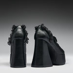A pair of black platform high heels with dramatic ruffled detailing on the sides, shown from the back against a dark gray gradient studio background.