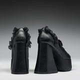 A pair of black platform high heels with dramatic ruffled detailing on the sides, shown from the back against a dark gray gradient studio background.
