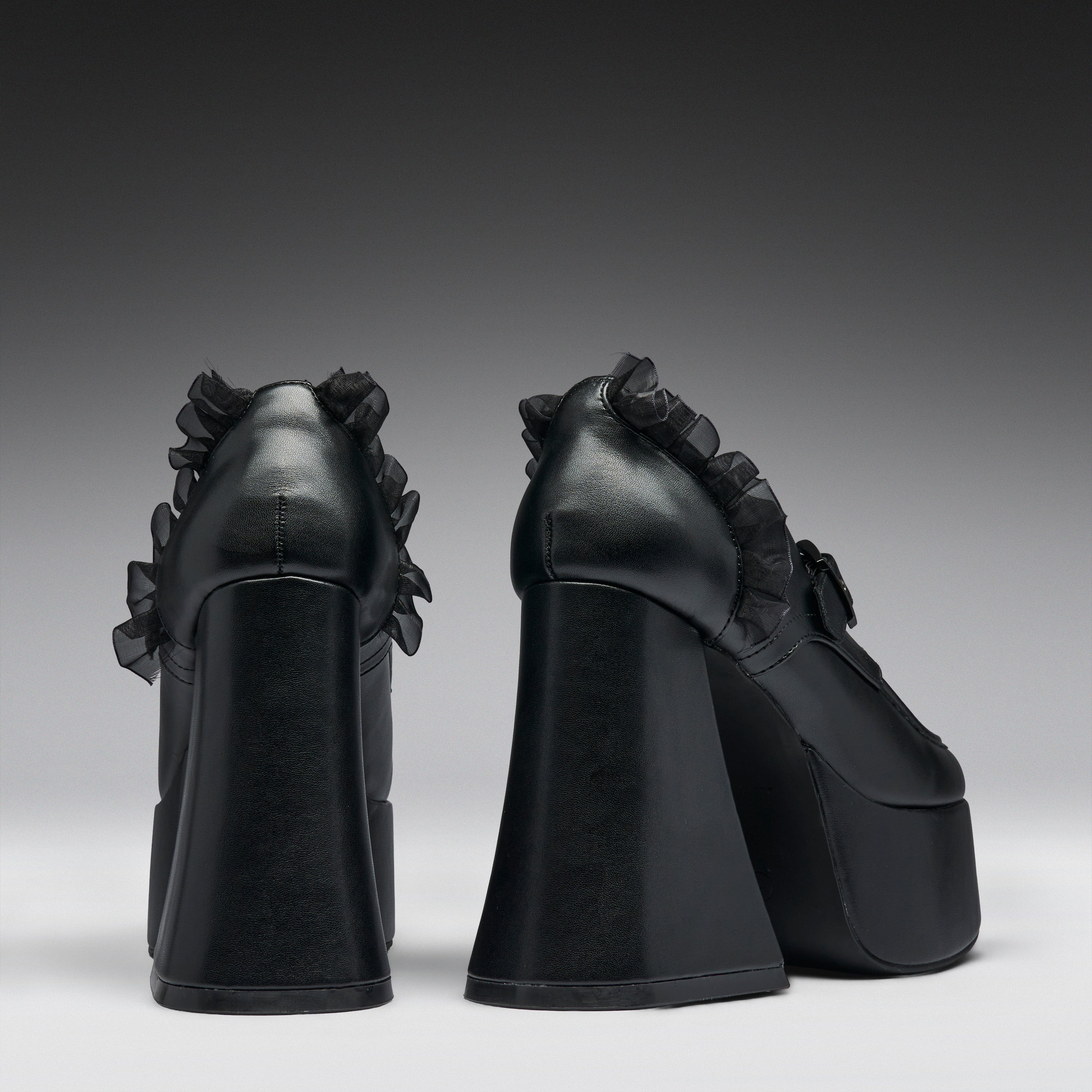 A pair of black platform high heels with dramatic ruffled detailing on the sides, shown from the back against a dark gray gradient studio background.