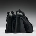 A pair of black platform high heels with dramatic ruffled trim along the sides, photographed from the back against a dark gray gradient studio background.