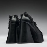 A pair of black platform high heels with dramatic ruffled trim along the sides, photographed from the back against a dark gray gradient studio background.