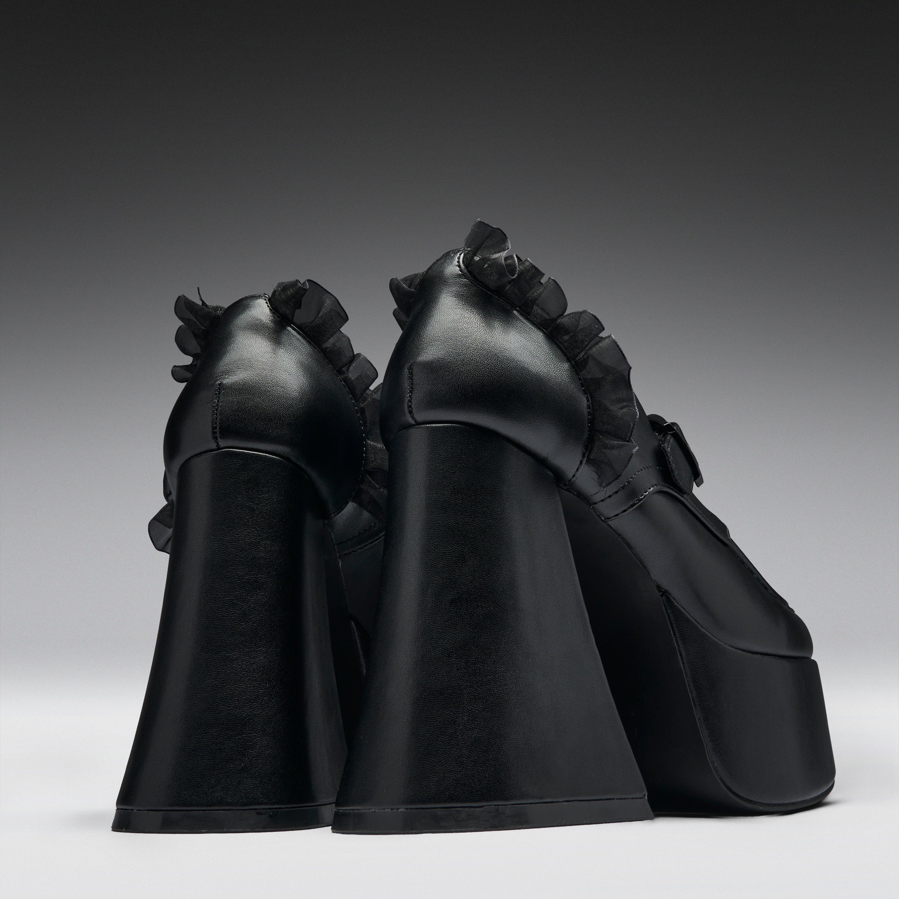 A pair of black platform high heels with dramatic ruffled trim along the sides, photographed from the back against a dark gray gradient studio background.