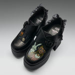 A pair of black platform Mary Jane-style shoes with ruffled trim and block heels, featuring printed character artwork on the toe boxes. The shoes are photographed on a clean light gray studio background.
