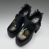 A pair of black platform Mary Jane-style shoes with ruffled trim and block heels, featuring printed character artwork on the toe boxes. The shoes are photographed on a clean light gray studio background.
