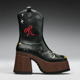 A black leather cowboy boot with a chunky brown wooden block heel and platform sole, decorated with embroidered stars, bows, and lightning bolts in red, yellow, and white.